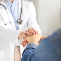 Lekarz w białym kitlu trzyma dłoń pacjenta, oferując wsparcie. W tle stetoskop.