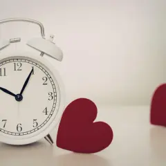 Biały budzik pokazuje 10:08. Obok niego leżą dwa czerwone serca. 0606 znaczenie: czas na miłość.