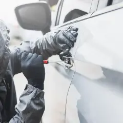 Osoba w kapturze i rękawiczkach próbuje otworzyć auto śrubokrętem. Sen o kradzieży auta może symbolizować utratę kontroli.