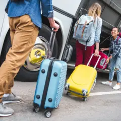 Ludzie z walizkami przy autobusie. Jedna walizka jest niebieska, druga żółta. W tle widać bagażnik autobusu.