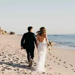 Para nowożeńców spaceruje po plaży. Panna młoda w pięknej sukni ślubnej z bukietem kwiatów, pan młody w granatowym garniturze. Słońce zachodzi nad oceanem.