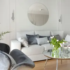 Przytulny salon z sofą i fotelami na tle jasnych, **białe ściany** zdobionych sztukaterią. Okrągłe lustro i bukiet tulipanów dodają elegancji.