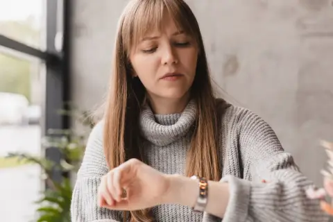 Zamyślona kobieta w szarym swetrze zerka na zegarek. Czyżby randka z aplikacją? 20 20 znaczenie dla singli.