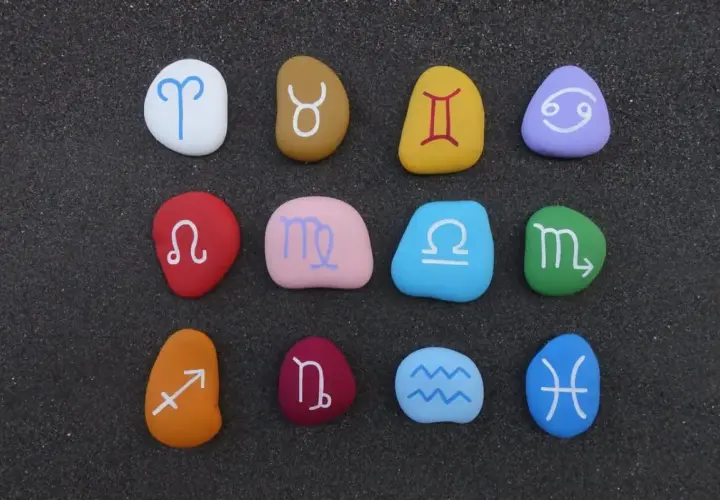 Kolorowe kamienie z symbolami znaków zodiaku po kolei, ułożone na ciemnym piasku.