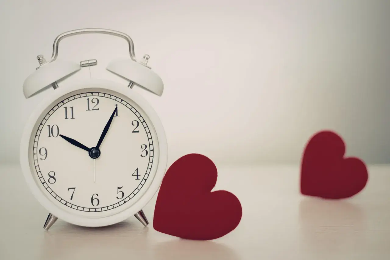 Biały budzik pokazuje 10:08. Obok niego leżą dwa czerwone serca. 0606 znaczenie: czas na miłość.
