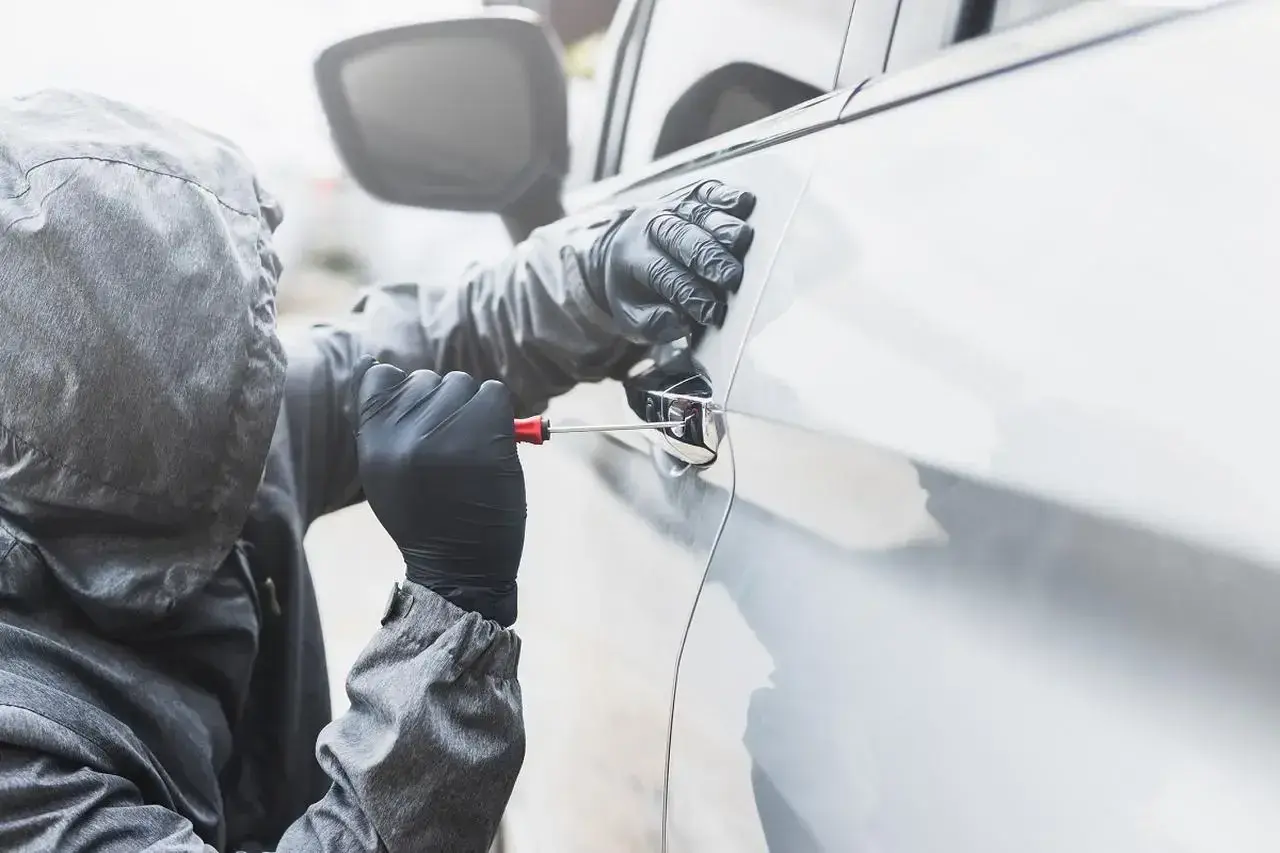 Osoba w kapturze i rękawiczkach próbuje otworzyć auto śrubokrętem. Sen o kradzieży auta może symbolizować utratę kontroli.