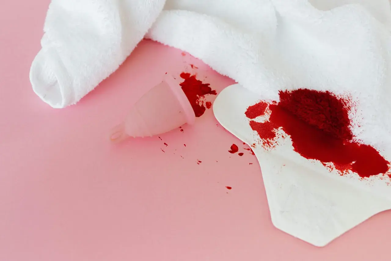 Sennik krew menstruacyjna: kubeczek i podpaska z plamami krwi na różowym tle.
