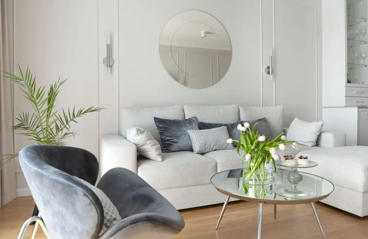 Przytulny salon z sofą i fotelami na tle jasnych, **białe ściany** zdobionych sztukaterią. Okrągłe lustro i bukiet tulipanów dodają elegancji.