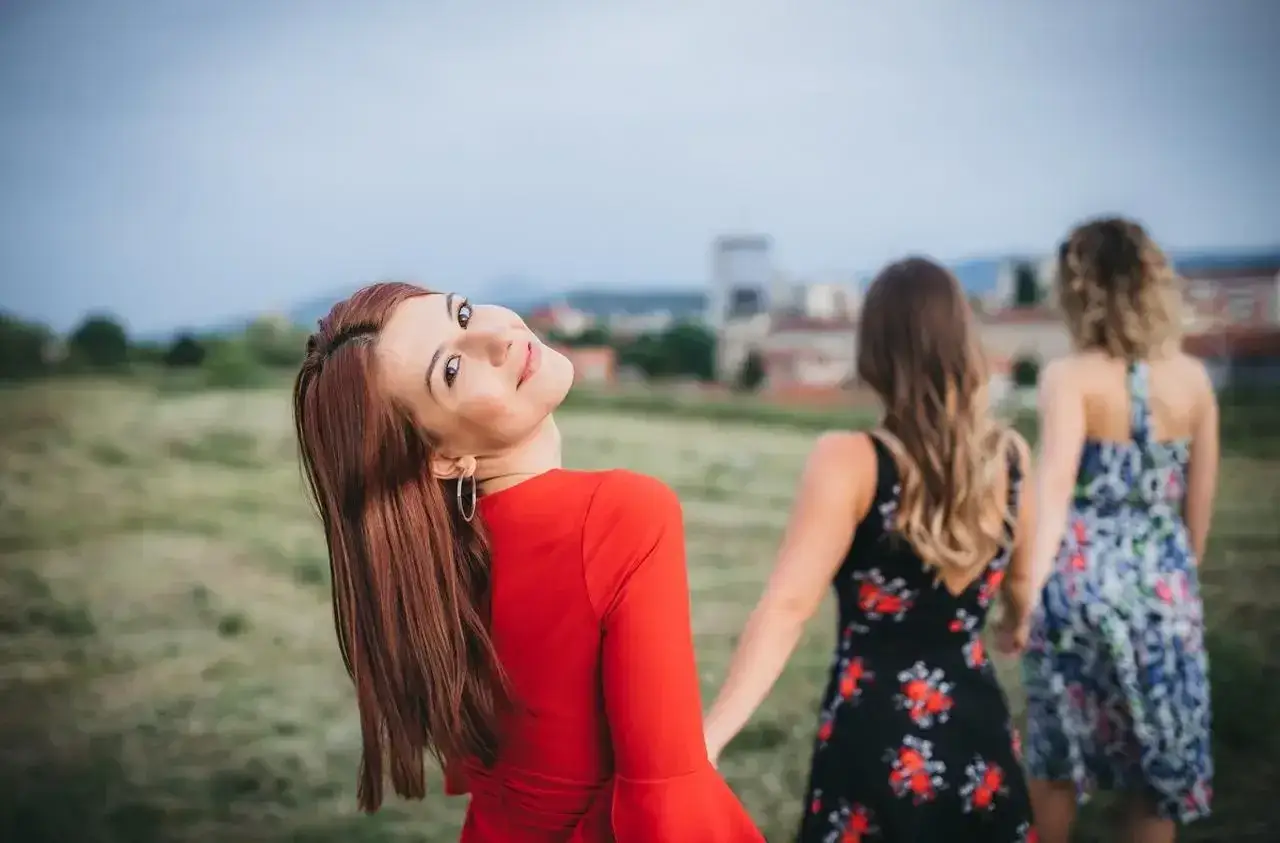 Uśmiechnięta kobieta w czerwonej sukience odwraca się do widza, podczas gdy dwie inne kobiety w sukienkach w kwiaty idą przed nią. Jak ze snu.