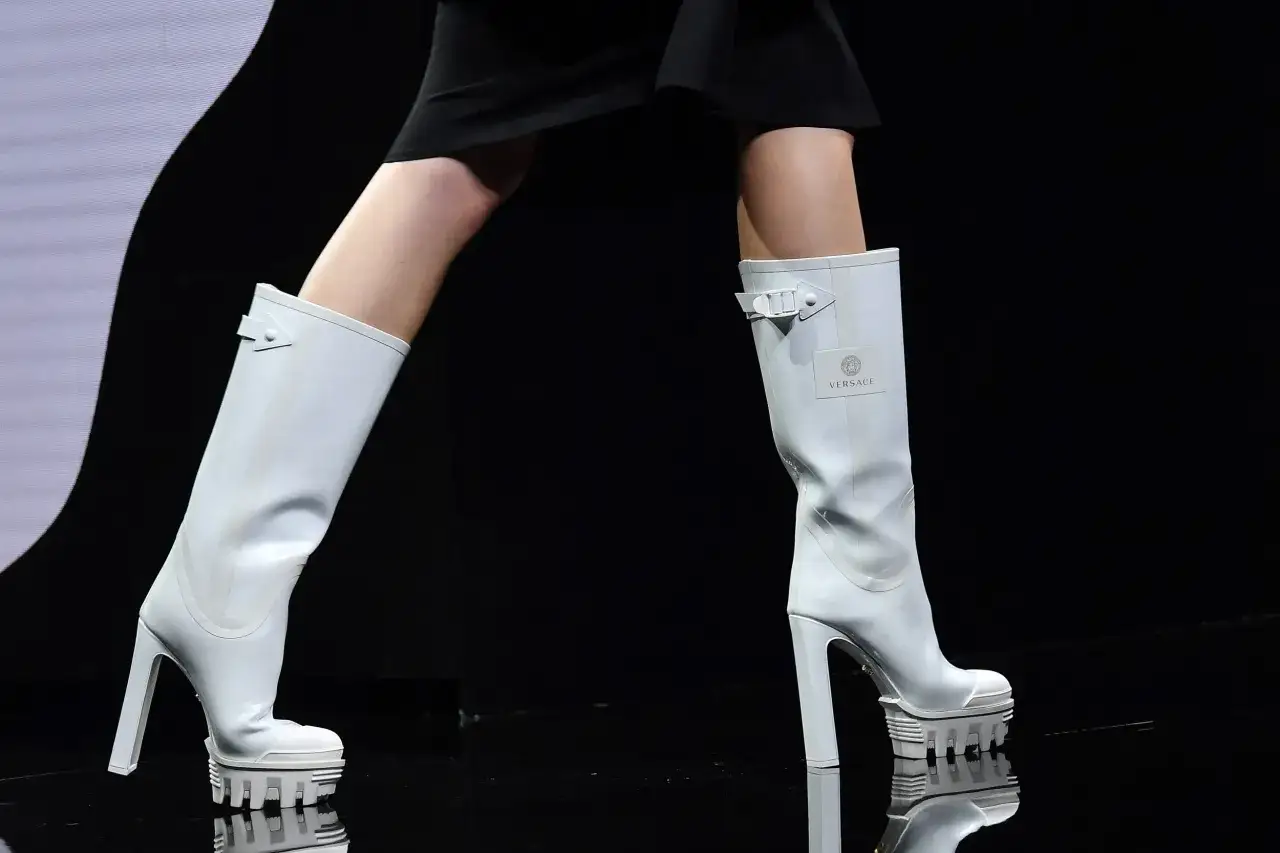 Białe, wysokie buty na obcasie z logo Versace. Wyglądają jak z sennika, dużo butów na wybiegu.